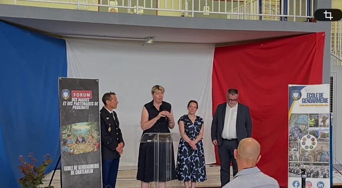 Quatre personnes debout sur une scène devant un drapeau tricolore français, lors d’un événement officiel. À gauche, un militaire en uniforme se tient à côté d’un panneau « Forum des maires et des partenaires de proximité, École de gendarmerie de Châteaulin ». Au centre, une femme s’exprime au micro, accompagnée d’une autre femme et d’un homme en costume. À droite se trouve un second panneau aux couleurs de l’école de gendarmerie, illustré de photos liées à la gendarmerie. Quelques personnes assises sont visibles au premier plan.