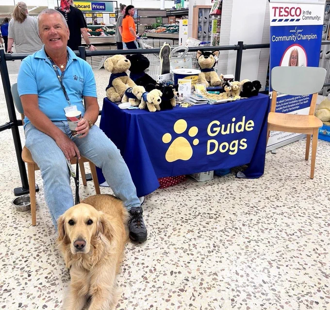 Photo shows a male volunteer sitting beside a GUIDE dogs trading stall with a golden retriever guide dog at his feet