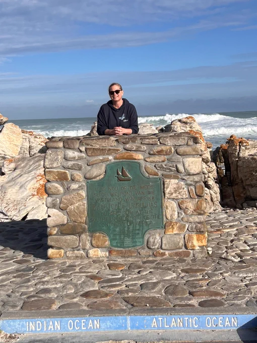 RES Managing Editor Jayne Whiffin in South Africa wearing a RES Large Blue butterfly mascot hoodie, standing behind a stone monument marking the division between the Indian Ocean and Atlantic Ocean with waves crashing in the background.