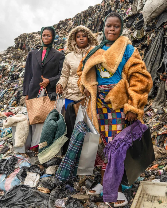 Three individuals stand in front of a large pile of waste, holding various bags and textiles. The background features stacked debris with clothing and other materials. The scene depicts a challenging environment with cloudy skies.
