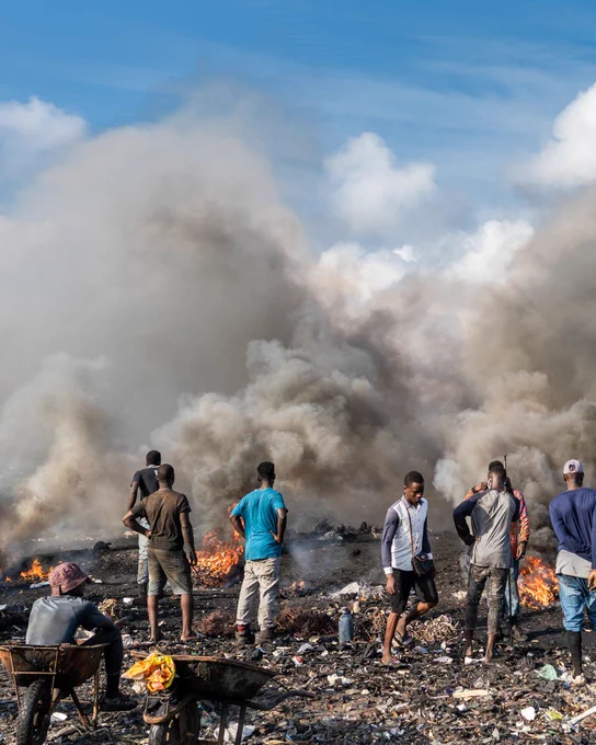 A group of people stands amidst a landscape filled with burning debris, producing thick smoke under a clear blue sky. The scene depicts an open-air waste site with various small fires scattered across the area.