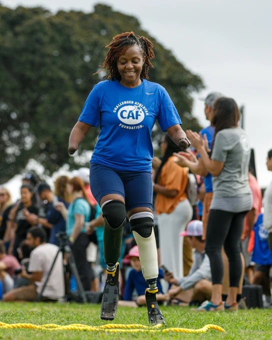 Smiling woman with limb differences runs on prosthetic legs at an outdoor event, wearing a blue Challenged Athletes Foundation shirt.