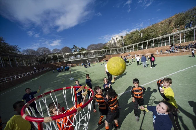 Niños jugando en cancha al aire libre, lanzando una gran pelota amarilla hacia un aro mientras otros miran desde las gradas.