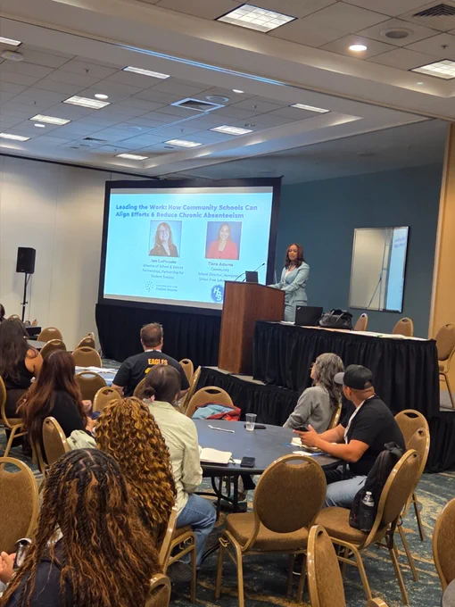 A close up shot of a crowded conference room, filled with attendees listening intently to Tiara behind the podium at the front of the room. A slide show is to the left with the header "Leading the Work: How Community Schools Can Align Efforts & Reduce Chronic Absenteeism" and two side by side headshots of Jen LoPiccolo and Tiara Adams.