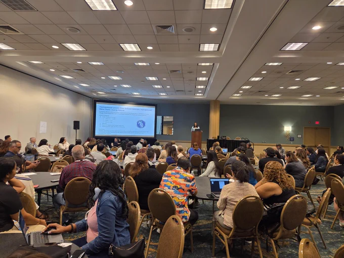 A crowded conference room, filled with attendees listening intently to Tiara behind the podium at the front of the room. A slide show is to the left with the header "Hempstead Union Free School District, Hempstead, New york - Long Island" and an Hempstead Union Free School District logo.