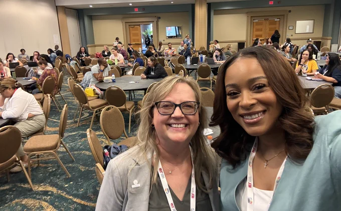 A selfie with Jen and Tiara with a crowded conference room behind them, filled with attendees sitting at circular tables. 