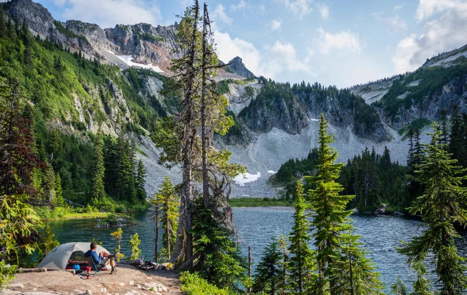 A camper relaxes in a chair next to a tent on the shore of Snow Lake, surrounded by dense evergreen forest and dramatic mountain cliffs in Mount Rainier National Park. Patches of snow remain on the rocky slopes under a partly cloudy sky. Photo by Evan Sloyka.