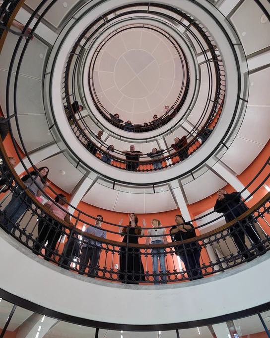 A group of people stand on multiple levels of a circular balcony inside a building. The view is looking up through the concentric levels, showcasing the architectural design and symmetry of the space.