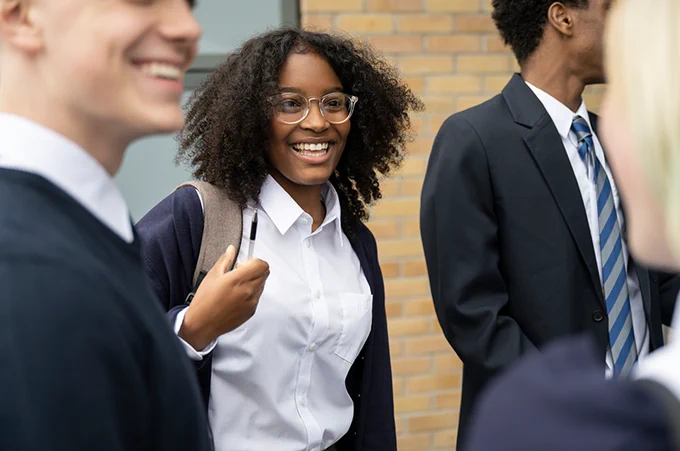 A group of people, dressed in school uniforms, are smiling and talking outside. One person is wearing glasses and carrying a backpack.