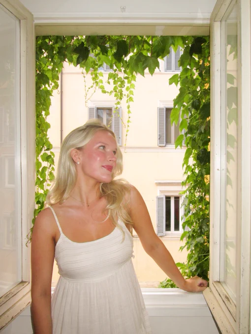 A person stands by an open window framed with green ivy, looking to the side. In the background, a building with several windows is visible.