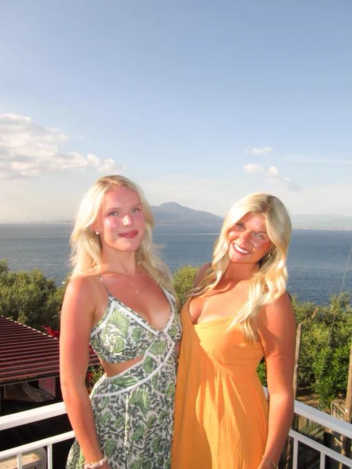 Two individuals standing on a balcony with a scenic view of the sea and mountains in the background. One person is wearing a green and white dress, and the other is dressed in an orange outfit. The sky is clear with some clouds.