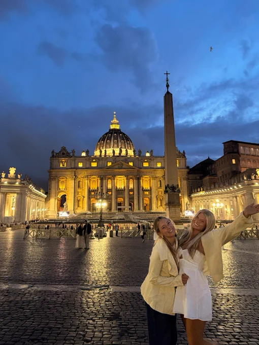 Two people standing in St. Peter's Square at dusk with St. Peter's Basilica lit up in the background. One person gestures upward, and the square is illuminated by lights, contrasting with the evening sky.