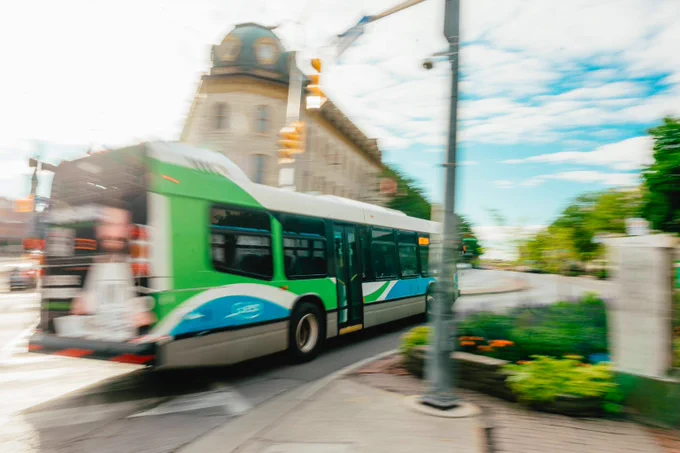Green and white city bus driving past a historic building on a sunny day with a partly cloudy sky.