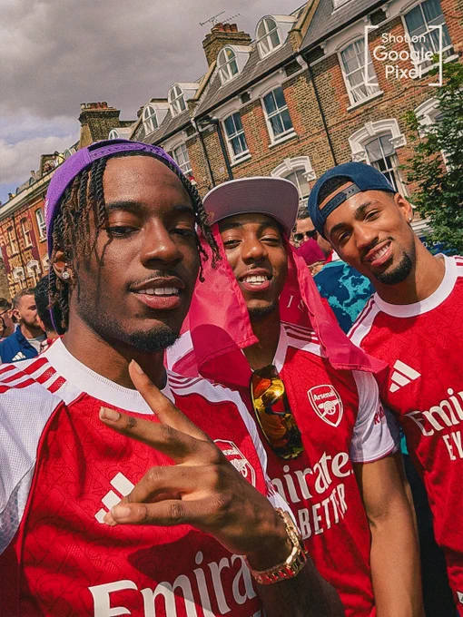 A selfie of three Arsenal players posing during the victory parade in front of classic London brick terraced houses. Noni in the foreground flashes a peace sign, while his teammates, Myles and Ethan, smile alongside him in their red and white kits. A "Shot on Google Pixel" watermark is in the top right corner.