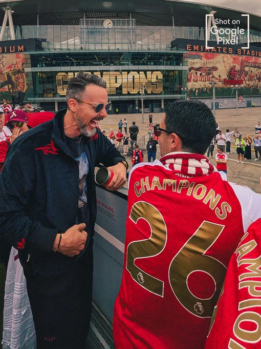 A candid shot outside the Emirates Stadium showing Josh Kroenke in a dark Arsenal jacket laughing heartily with Mikel Arteta. Mikel's back is turned, showing an Arsenal shirt printed with "CHAMPIONS 26" in bold gold lettering. In the background, the massive gold word "CHAMPIONS" spans the stadium facade. A "Shot on Google Pixel" watermark is in the top right corner.
