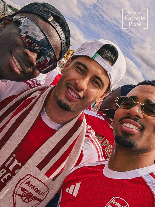 A close-up, high-energy selfie of Arsenal players Bukayo Saka, Gabriel Martinelli and Gabriel Jesus celebrating outdoors. Martinelli is in the center wearing a backward white cap and an Arsenal scarf, while Jesus smiles on the right in sunglasses. A "Shot on Google Pixel" watermark is in the top right corner.