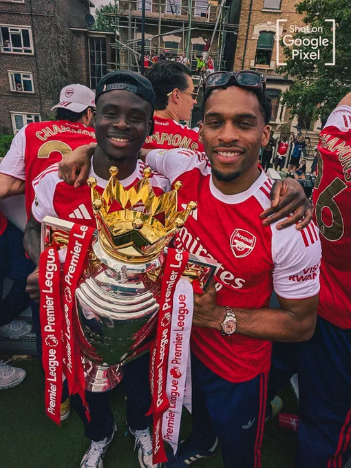 A joyful medium shot of Bukayo Saka and Jurriën Timber standing close together and smiling brightly as they hold up the Premier League trophy. The trophy features its iconic golden crown and is draped in red and white ribbons. A "Shot on Google Pixel" watermark is in the top right corner.