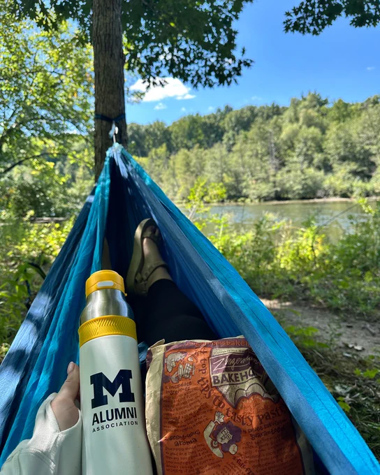A person lies in a hammock in front of a forest of green trees and a river. They're holding a Michigan alumni water bottle, plus a bag from Zingerman's Bakehouse.