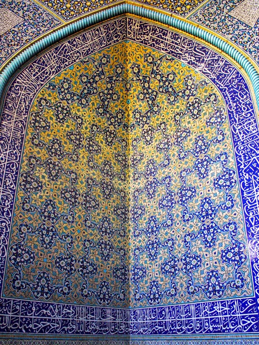A close-up, capturing the intricate tilework in a corner convergence within the interior prayer chamber of the Sheikh Lotfollah Mosque in Isfahan, Iran. The image shows two intersecting walls under a curved arch, completely covered in vibrant Safavid-era glazed mosaic tiles. The central patterns feature incredibly detailed floral arabesques and repeating botanical motifs in shades of cobalt blue, turquoise, creamy white, and saffron yellow. Running along the bottom and top borders of the structure are horizontal panels filled with elegant, fluid Islamic calligraphy in white Thuluth script set against a deep blue background.
