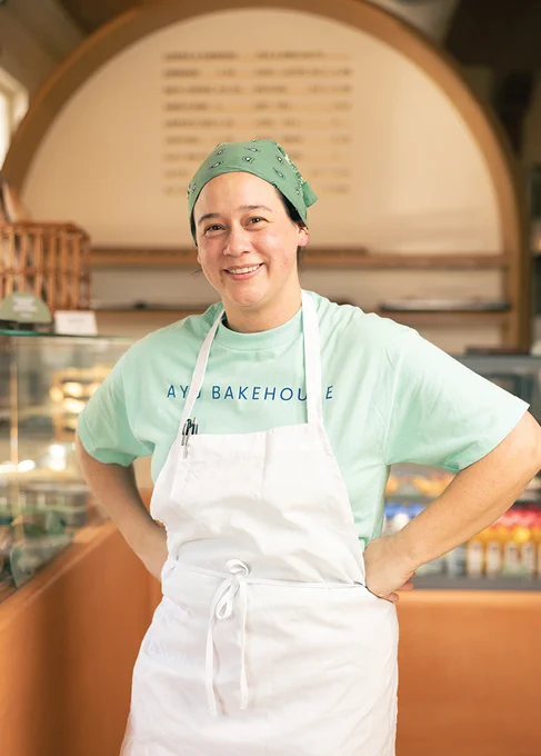 A person wearing a green bandana, light green T-shirt with "Ayu Bakehouse" text, and a white apron stands inside a bakery. The background shows a menu board, shelves, and some snacks on display.
