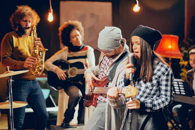 Group of four musicians playing saxophone, guitar, bass, and singing in a cozy, warmly lit room with hanging bulbs and a red lamp.