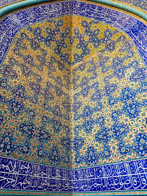A close-up, capturing the intricate tilework in a corner convergence within the interior prayer chamber of the Sheikh Lotfollah Mosque in Isfahan, Iran. The image shows two intersecting walls under a curved arch, completely covered in vibrant Safavid-era glazed mosaic tiles. The central patterns feature incredibly detailed floral arabesques and repeating botanical motifs in shades of cobalt blue, turquoise, creamy white, and saffron yellow. Running along the bottom and top borders of the structure are horizontal panels filled with elegant, fluid Islamic calligraphy in white Thuluth script set against a deep blue background.