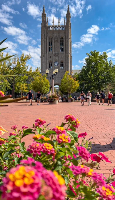 Tall gothic-style clock tower rising above a bustling plaza with people walking and colorful flowers in the foreground under a partly cloudy sky by Janie Adams.