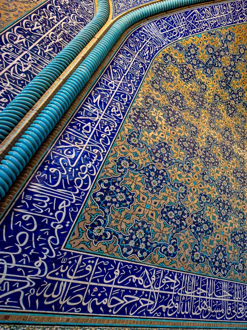 A close-up, capturing the intricate tilework in a corner convergence within the interior prayer chamber of the Sheikh Lotfollah Mosque in Isfahan, Iran. The image shows two intersecting walls under a curved arch, completely covered in vibrant Safavid-era glazed mosaic tiles. The central patterns feature incredibly detailed floral arabesques and repeating botanical motifs in shades of cobalt blue, turquoise, creamy white, and saffron yellow. Running along the bottom and top borders of the structure are horizontal panels filled with elegant, fluid Islamic calligraphy in white Thuluth script set against a deep blue background.