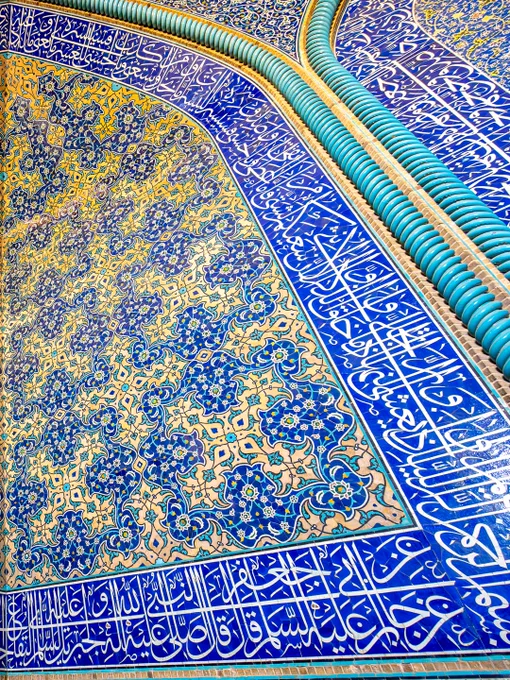 A close-up, capturing the intricate tilework in a corner convergence within the interior prayer chamber of the Sheikh Lotfollah Mosque in Isfahan, Iran. The image shows two intersecting walls under a curved arch, completely covered in vibrant Safavid-era glazed mosaic tiles. The central patterns feature incredibly detailed floral arabesques and repeating botanical motifs in shades of cobalt blue, turquoise, creamy white, and saffron yellow. Running along the bottom and top borders of the structure are horizontal panels filled with elegant, fluid Islamic calligraphy in white Thuluth script set against a deep blue background.