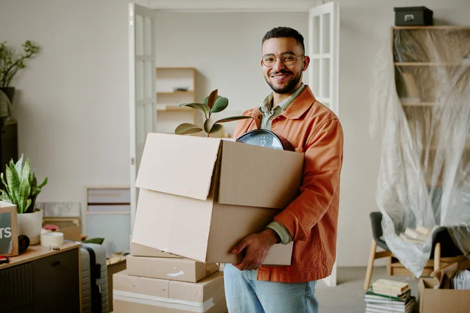 Person carrying a cardboard box filled with household items, including a plant and a clock, in a room with packed boxes and furniture.