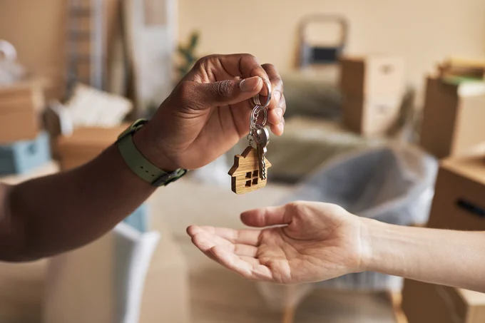 Hand passing a house-shaped keychain with keys to another person in a room with moving boxes.