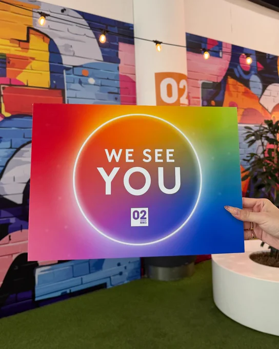 The image shows a hand holding a colourful poster with the text "WE SEE YOU" and "O2" in white. The background features a vibrant, abstract mural with various colours and geometric shapes, illuminated by string lights.