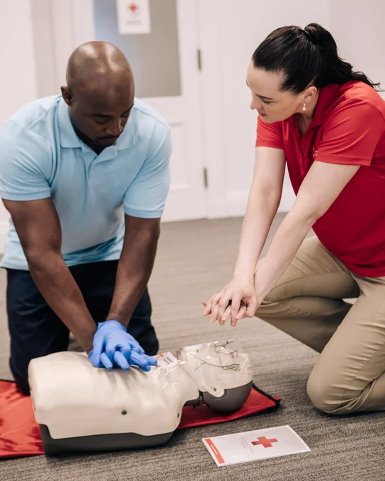 Two individuals practice CPR on a mannequin in a room with a Red Cross sign. One person is demonstrating chest compressions, while the other observes. A Red Cross booklet is on the floor nearby.