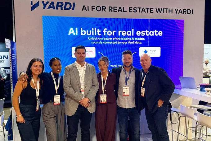 A group of six people stand together in front of a large display for "Yardi AI for Real Estate," at an event. They are smiling and wearing event badges. The backdrop showcases text about AI models connected to Yardi data.