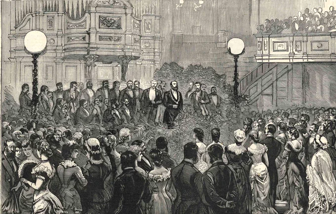 A formal gathering in a grand hall with a pipe organ, men on a decorated platform, and a large audience.