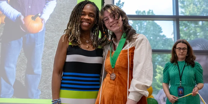 Two people standing indoors near a large window, one wearing a striped dress and the other an orange dress with a green medal around their neck.