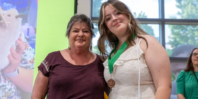 Two people standing side by side indoors, one wearing a white dress with a green medal around their neck, the other in a maroon shirt.