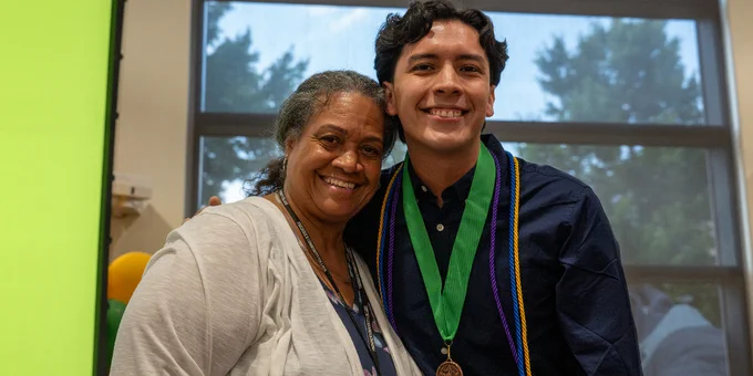 Two people standing closely together indoors near a large window with trees visible outside, one wearing a dark shirt and graduation cords.