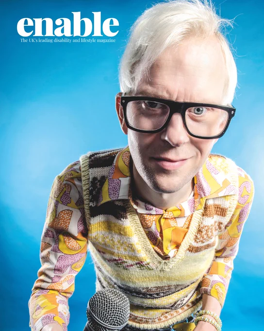 An image of Robert White. He is smiling, has white hair and is wearing glasses. He is holding a microphone and wearing a bright patterned shirt and yellow knitted cardigan.