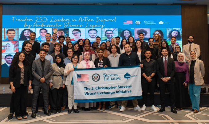 Posed photo of a group of young people. Behind them, a screen reads "Freedom 250: Leadership in Action Inspired by Ambassador Stevens' Legacy. Some of the young people are also holding a banner that reads "J. Christopher Stevens Virtual Exchange Initiative" with four logos (U.S. flag, State Department Seal, Stevens Initiative logo, Aspen Institute logo).