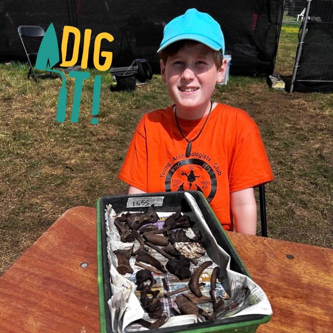 A young person wearing an orange t-shirt with the words 'Young Archaeologists Club' written on it is sitting at a wooden trestle table outside on a sunny day. On the table is a green tray lined with newspaper and filled with recently excavated pieces of animal bone. 