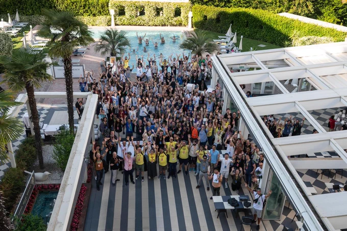 A large group of PyCon Italia attendees, volunteers, and organizers gathered outdoors near a pool, waving at the camera during the closing group photo of the conference.