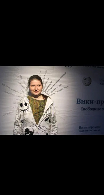 Photo of a Alexandra Elbakyan standing against a projected Russian-language backdrop referencing free knowledge, wearing a white patterned hoodie over a green top, with soft lighting and a shadowed presentation-style background.