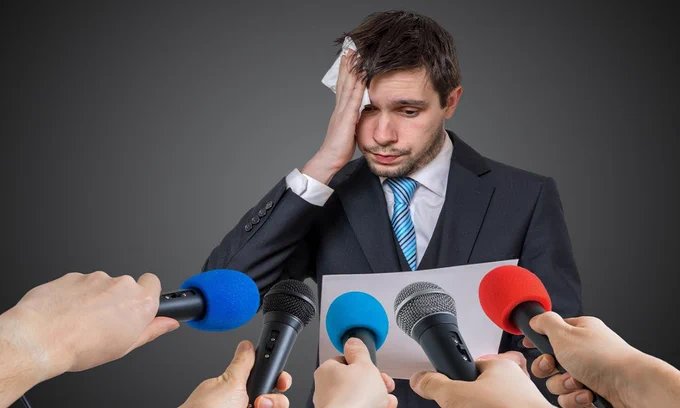 Man looking nervous with 5 microphones in front of him.