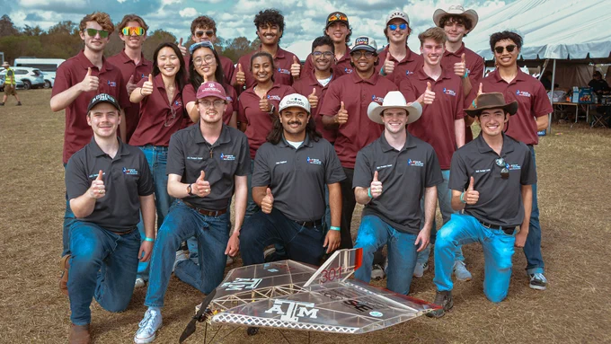 A group of individuals wearing maroon and gray shirts with the Texas A&M University logo stand on grassy ground, some giving thumbs up. In front, a model aircraft with Texas A&M insignia is displayed. A large tent and trees are visible in the background.