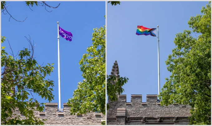 Two flags flying against a blue sky. On the left is the purple and white Haudenosaunee (Iroquois) flag. On the right is a Pride flag. 