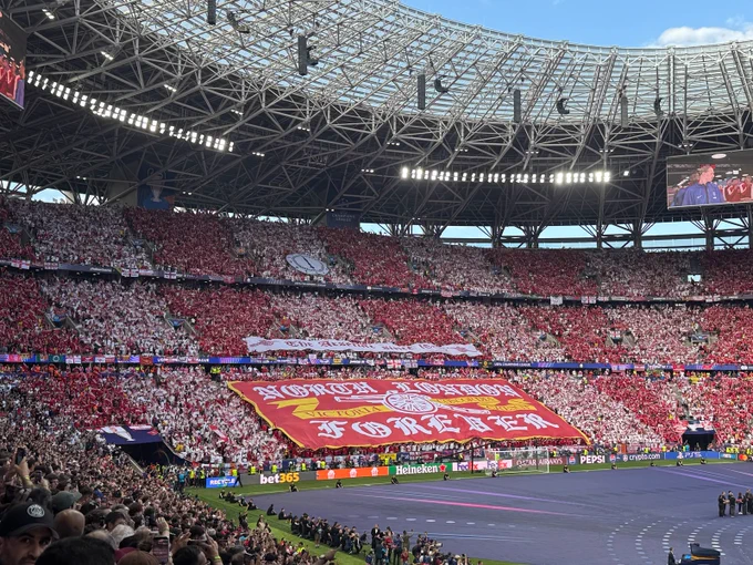 Supporters behind a giant red-and-white tifo displaying the words “North London Forever” during a packed UEFA Champions League final, with thousands of fans holding scarves aloft.