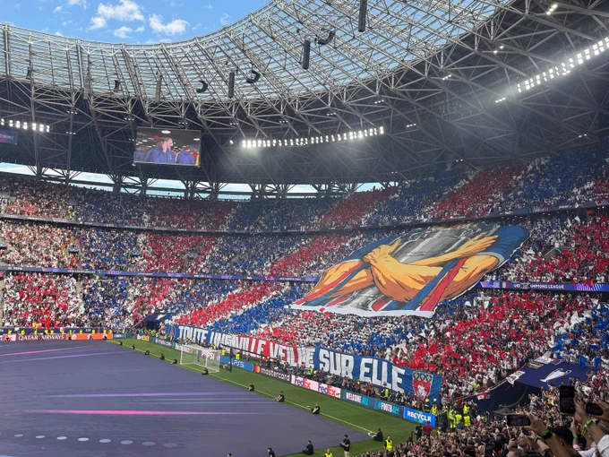 A large red, white and blue tifo featuring the UEFA Champions League trophy is displayed by supporters in a packed stadium before kick-off.