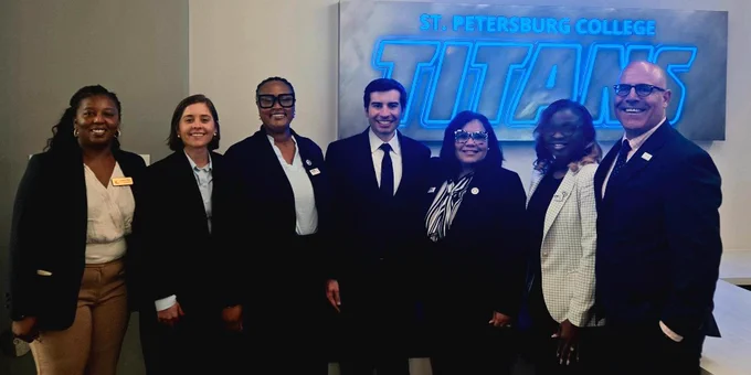 A group of six individuals from SPC leadership in formal attire stands next to Keith Sonderling in front of a sign that reads "St. Petersburg College Titans." They are smiling and positioned in an indoor setting with neutral-colored walls.