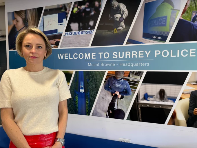 Image shows Commissioner Lisa Townsend who is a blonde, short haired woman standing to the left of the picture.  She is looking straight at the camera with a serious face and is wearing a cream coloured short sleeved woollen top and red skirt.  She is standing beside a large wall sign that says 'Welcome to Surrey Police', Mount Browne Headquarters.  The text is surrounded by different policing images 